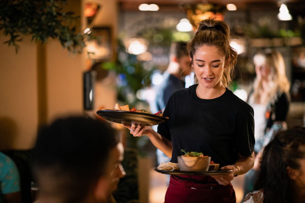 Kvinnlig servitris bär två tallrikar med mat i en restauranglokal. Förenklingsrådet föreslår att krav på matservering och eget kök för alkoholservering i restauranger tas bort.