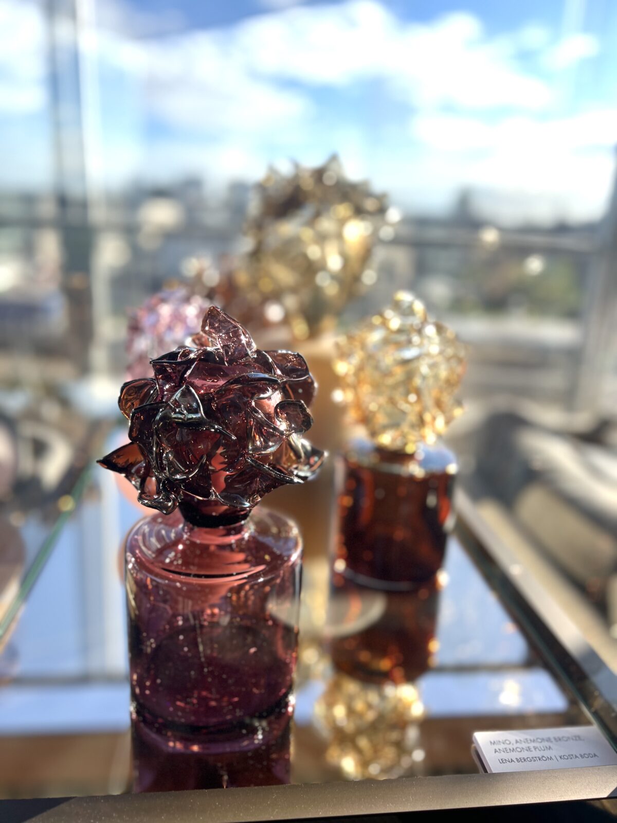 Lena Bergströms Mino, Anemone bronze, och Anemone plum tillverkad hos Kosta Boda gnistrar i en glasmonter mellan borden på terrassen.